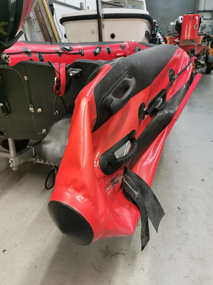 A red Rigid Inflatable Boat (RIB) in a workshop showing severe structural delamination, where the black rubber strake and tube fabric have completely detached from the transom and hull.