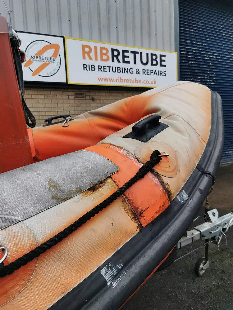 An orange RIB collar showing extreme UV damage, chalking, and "crazing" of the fabric surface, parked in front of the RIB Retube workshop in Livingston, Scotland.